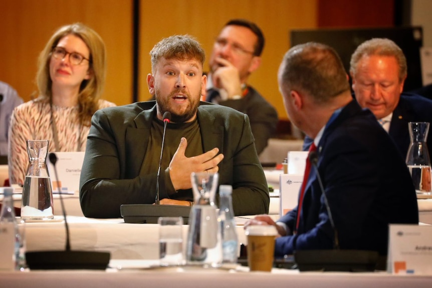 A mad in a suit jacket sits at a table speaking as business and union leaders look on.