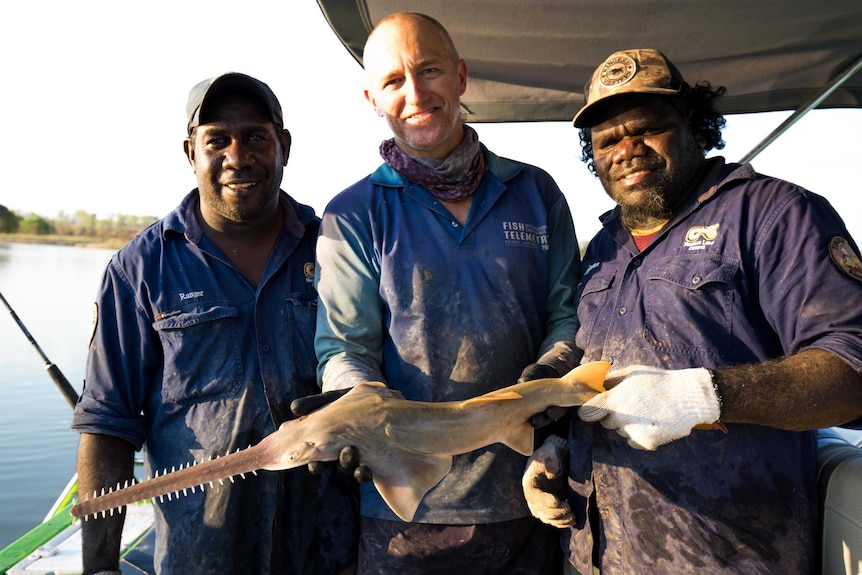 Three men hold a sawfish on a boat.
