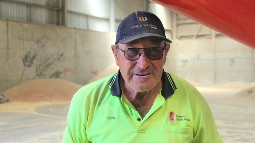 He wears a cap and stands in a shed with piles of grain