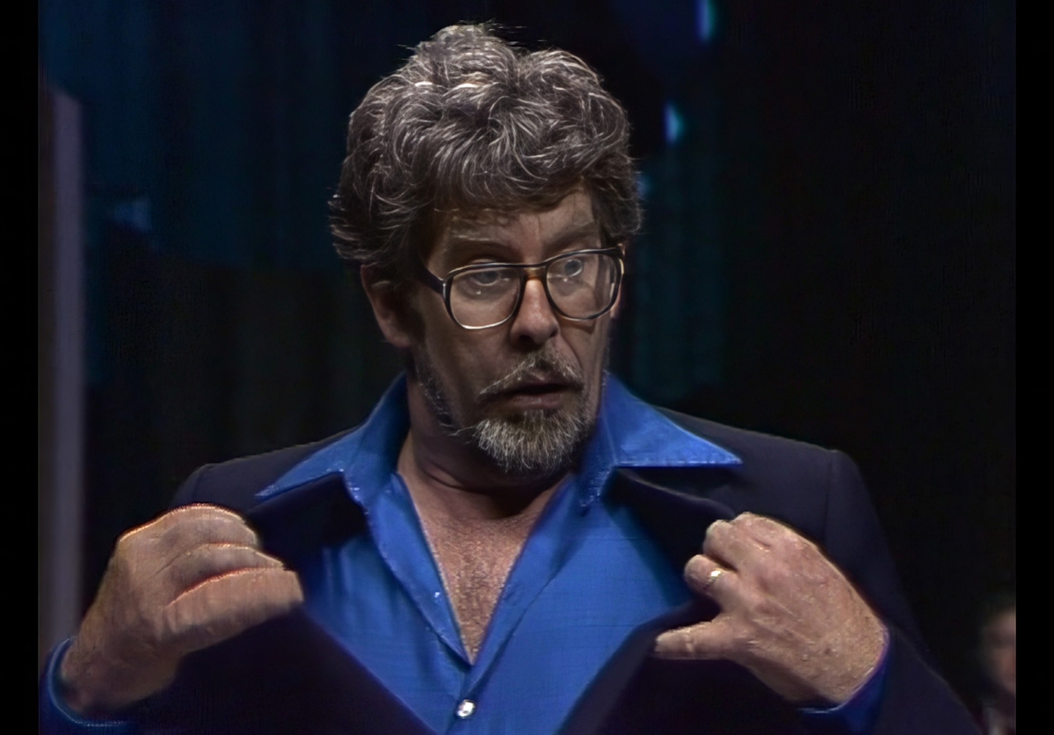 A man in a blue and black suit pulls at his collar with both hands.