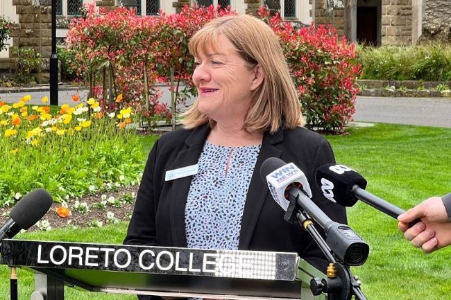 A middle-aged woman wearing a dark blazer stands outside speaking at a lectern.