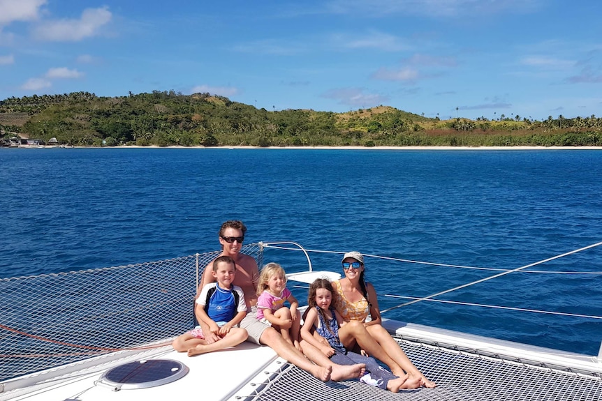 two adults and three children sit on a white yacht with the ocean and an island in the background