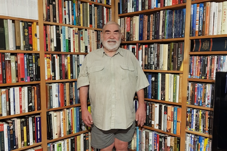 A man with short grey hair and a beard smiling while standing at the corner of several large, filled bookshelves.
