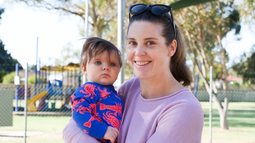 A woman holding a baby boy smiling and looking at the camera with a playground and trees in the background