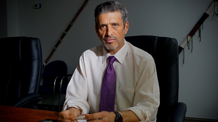 A man in a white shirt and purple tie sits at a table with a serious expression
