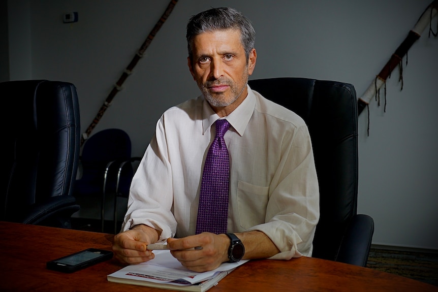 A man in a white shirt and purple tie sits at a table with a serious expression