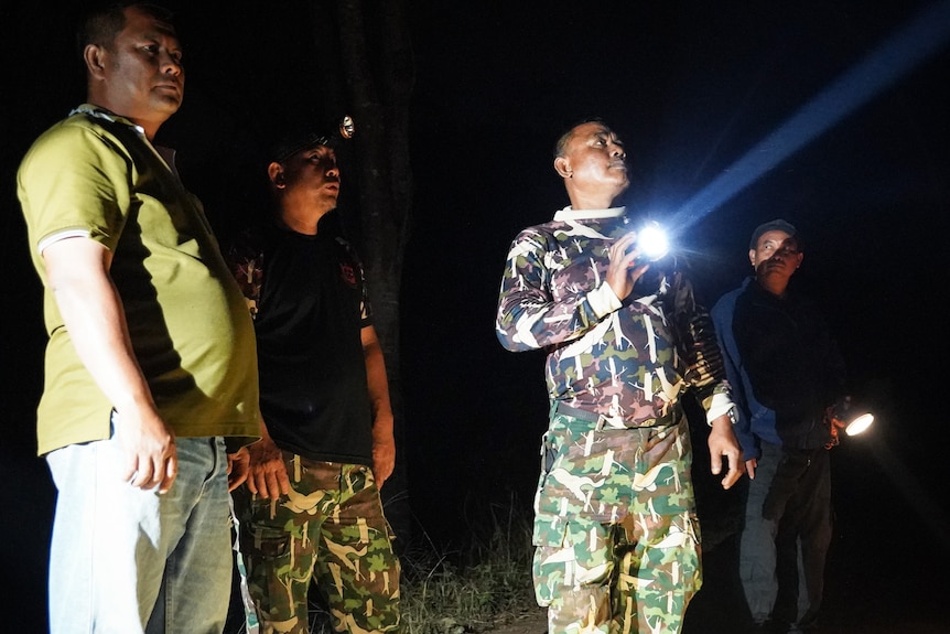 A man dressed in camoflage gear holds a flashlight and shines it on a field as three other men watch on.