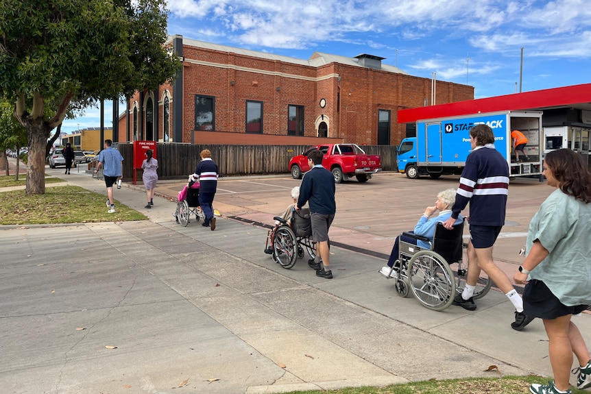 Three students push three ages care residents in their wheelchairs along a footpath.