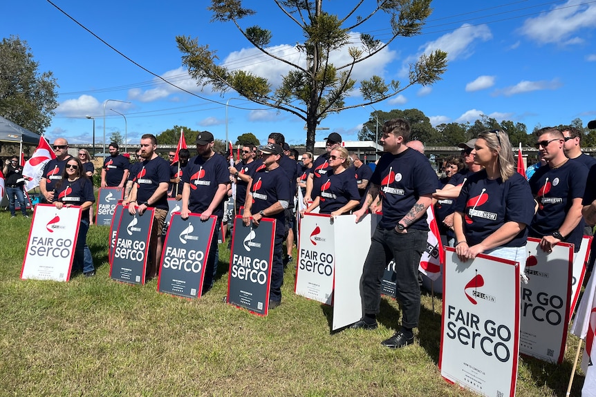 Group of prison officers wear union uniforms and carry signs reading "fair go Serco". 
