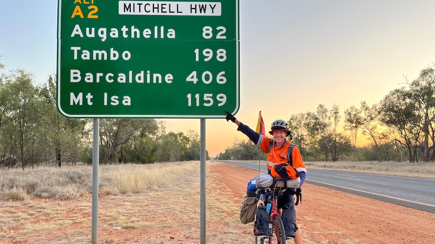 A man on a pushbike pointing at a sign that says Mount Isa 1159 kms