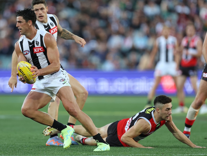 A Collingwood AFL player holds the ball and props to run away from a potential tackler left lying on the ground during a game.