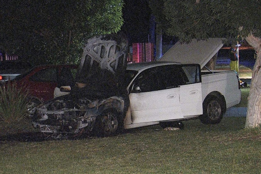 A burnt out white ute sits parked on the lawn of a house with its doors open.