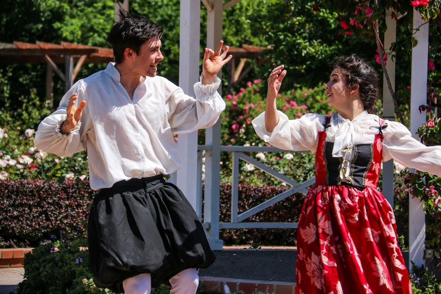 A male and female performer, wearing period clothing, gesture animatedly as they act in a park.