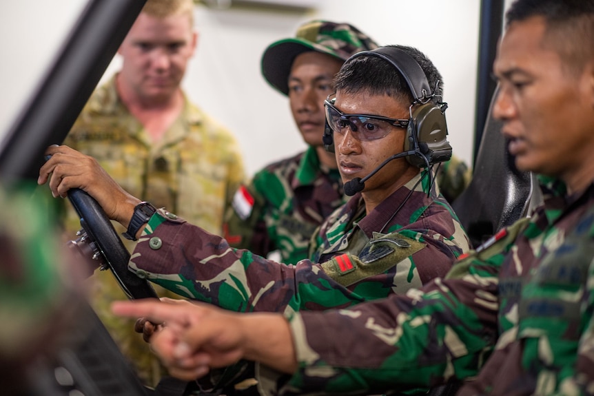 An Indonesian soldier at a steering wheel, while another nearby points and an Australia soldier in the background watches on.