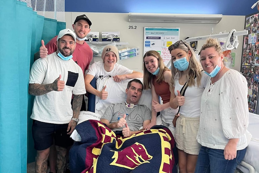 A group of people smiling with thumbs up around a man sitting up in hospital bed also doing thumbs up