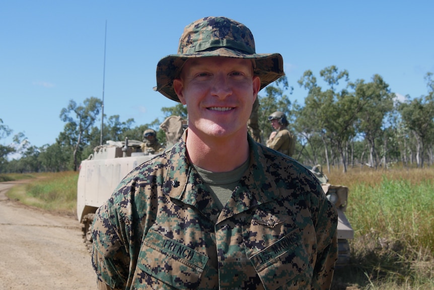 A man wearing an army green camouflage bucket hat and uniform, standing in front of a tank in bushland