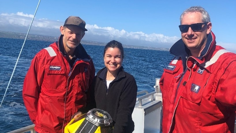 Three people stand on a boat. A woman smiles and holds a yellow bouy.