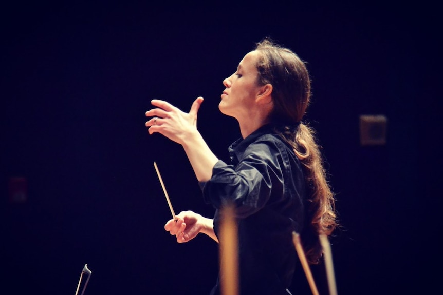 Close up of Jessica Cottis conducting the Sydney Symphony Orchestra against a completely black background.