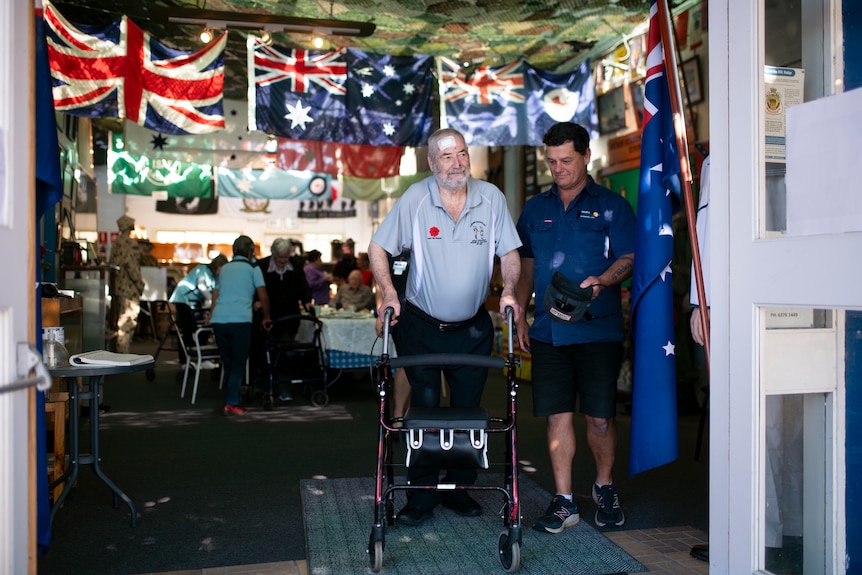 An older gentleman smiles as he exits a room filled with flags using a walker and guided by support staff.