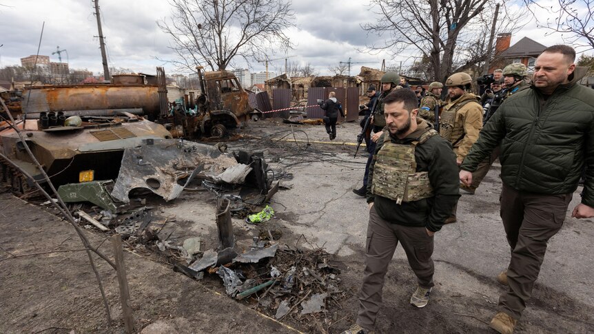 Volodymyr Zelenskyy walks past destroyed military vehicles followed by an entourage in Bucha