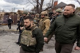 Volodymyr Zelenskyy walks past destroyed military vehicles followed by an entourage in Bucha