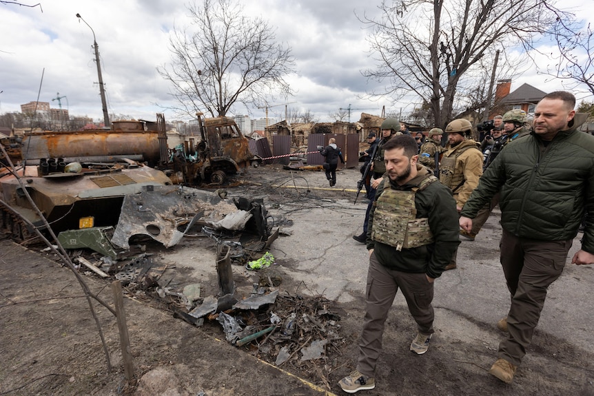 Volodymyr Zelenskyy walks past destroyed military vehicles followed by an entourage in Bucha