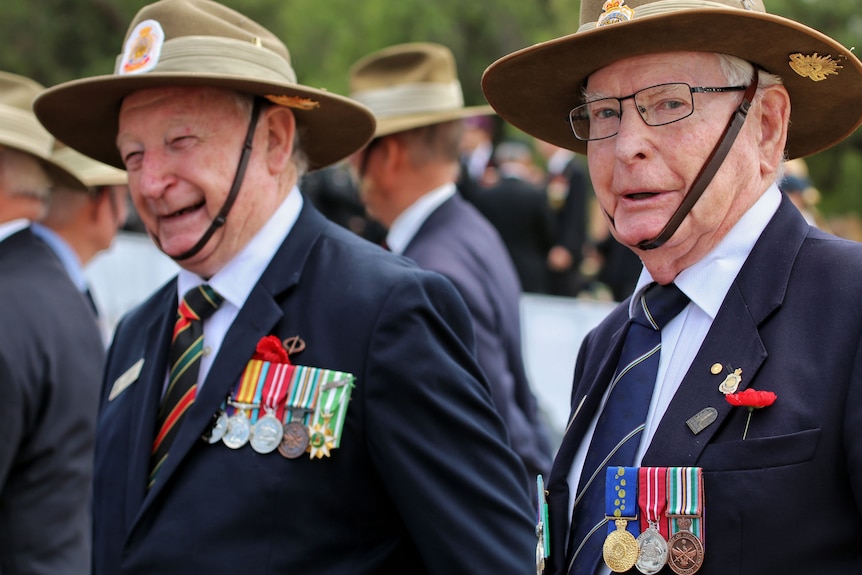 Two older men, both wearing uniforms with service badges, stand in a crowd of veterans. They are both smiling.