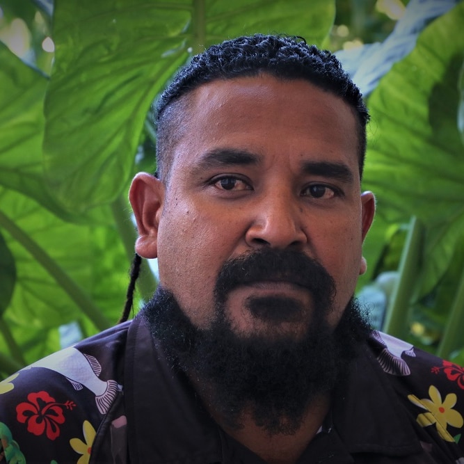 Close up of 37-year-old Torres Strait Islander man looking concerned with green plants in the background.