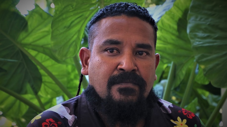 Close up of 37-year-old Torres Strait Islander man looking concerned with green plants in the background.