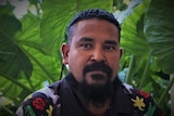 Close up of 37-year-old Torres Strait Islander man looking concerned with green plants in the background.