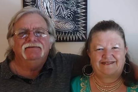 Man with glasses and grey hair and long mustache, smiling, next to a woman with long brown hair and large circle earrings