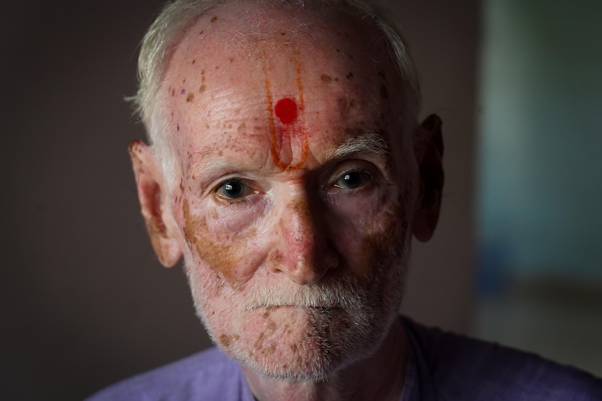 An older man with red marking on his forehead looks directly into camera