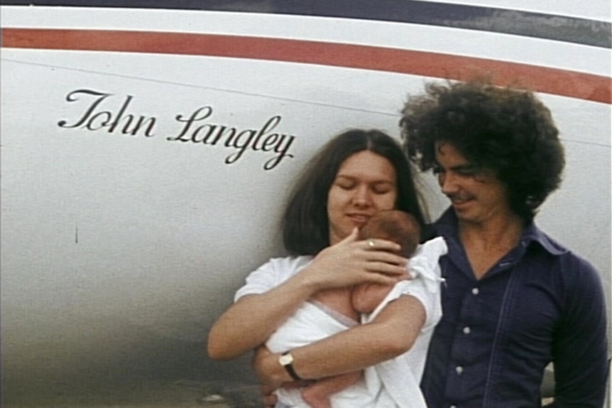 A young mother cradles her baby as the father looks on in front of an aircraft