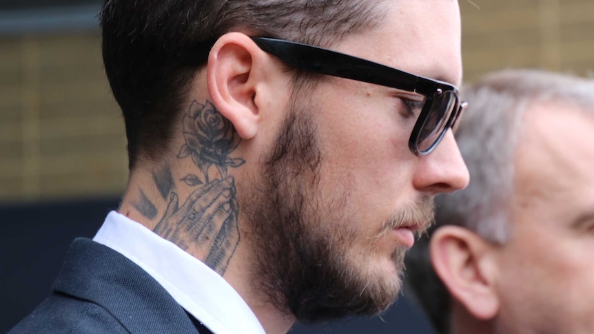 A man with a neck tattoo stands outside court wearing a suit and sunglasses.