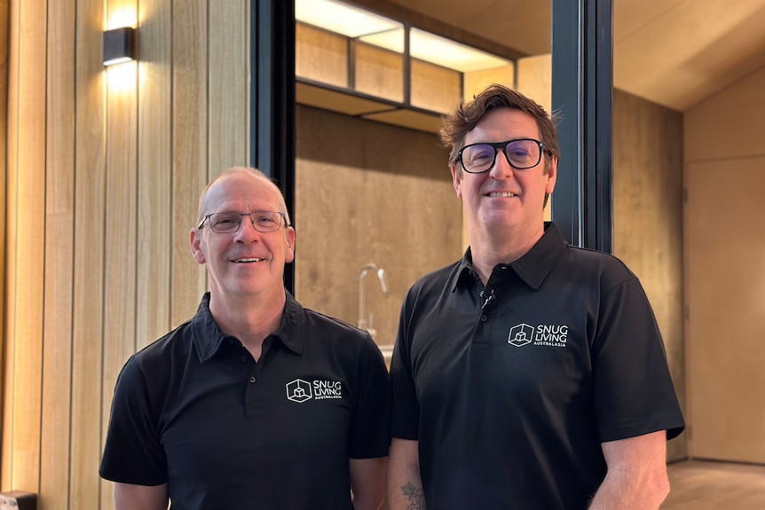Two men in black polo shirts and glasses smile at the camera. 