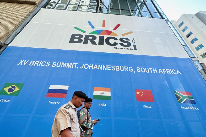People walk past a convention centre with a large billboard advertising the BRICS summit with flags of each country.