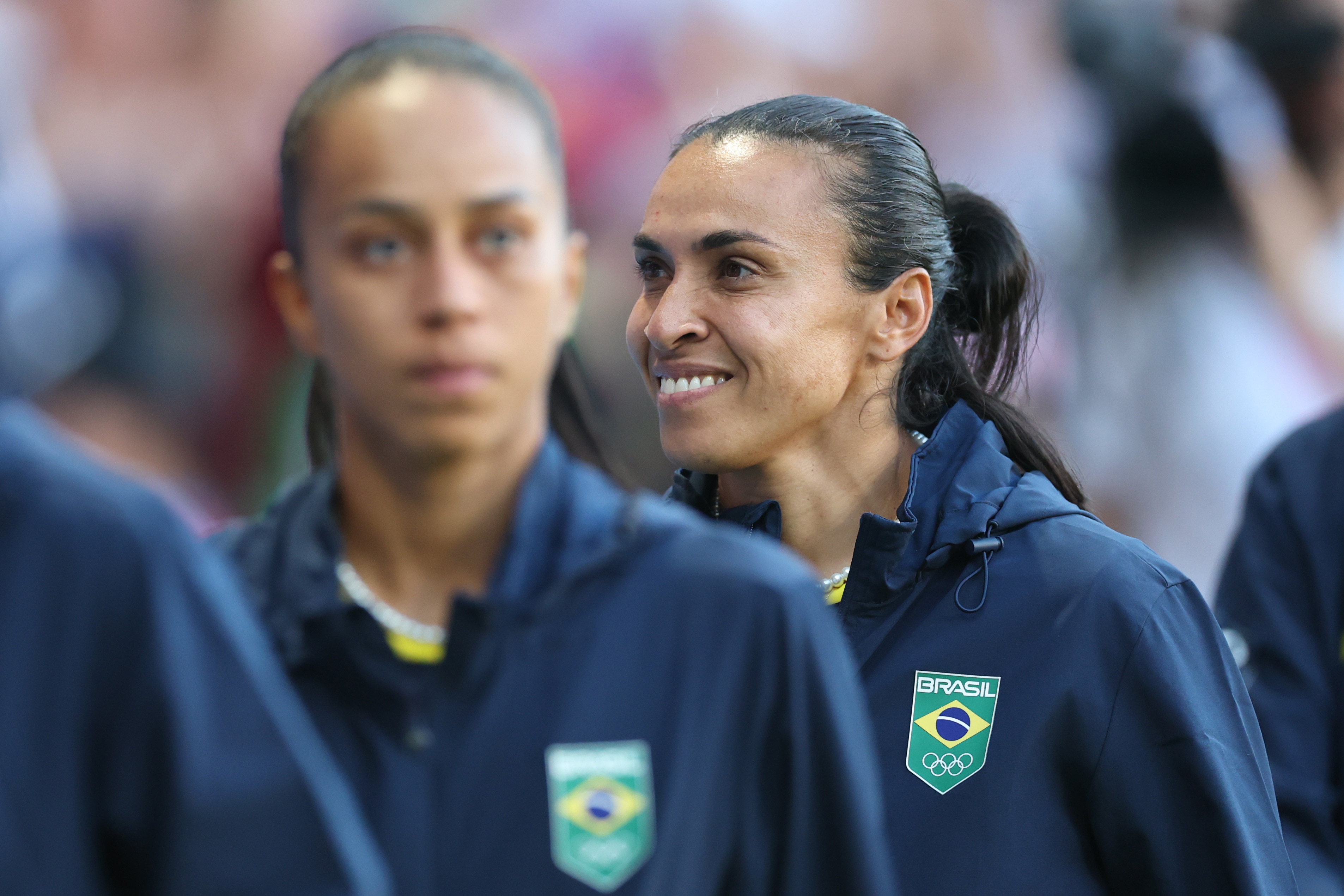 A woman wearing a blue jacket with the Brazil log smiles while standing behind a team-mate