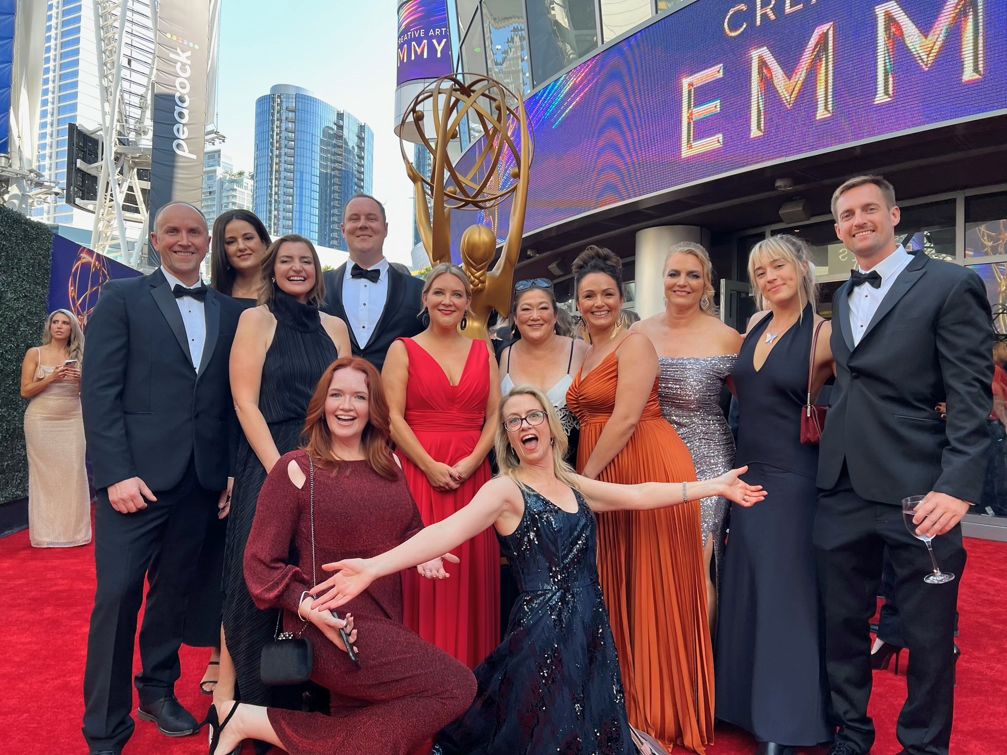 12 people pose for a photo at a red carpet. A giant Emmy statue is behind them.