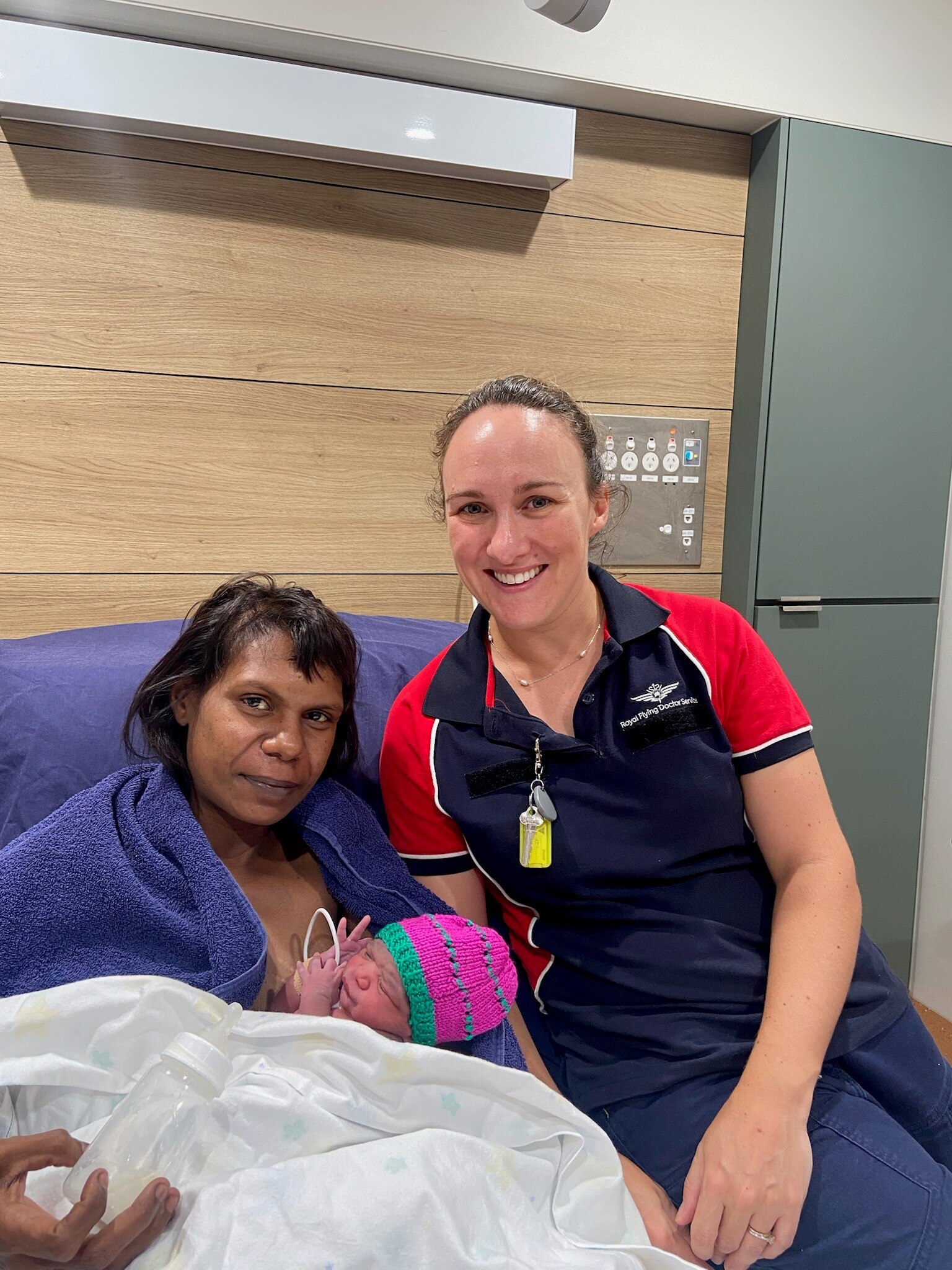 A woman with a baby on her breast sits in a hospital bed next to a smiling female nurse.