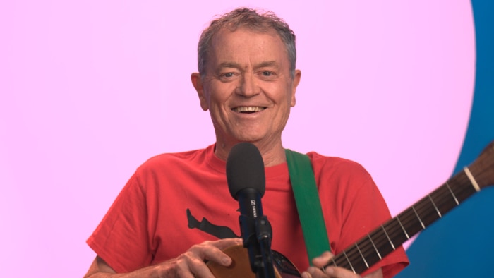A person smiling, playing a guitar in front of a microphone, with a pink and blue background.