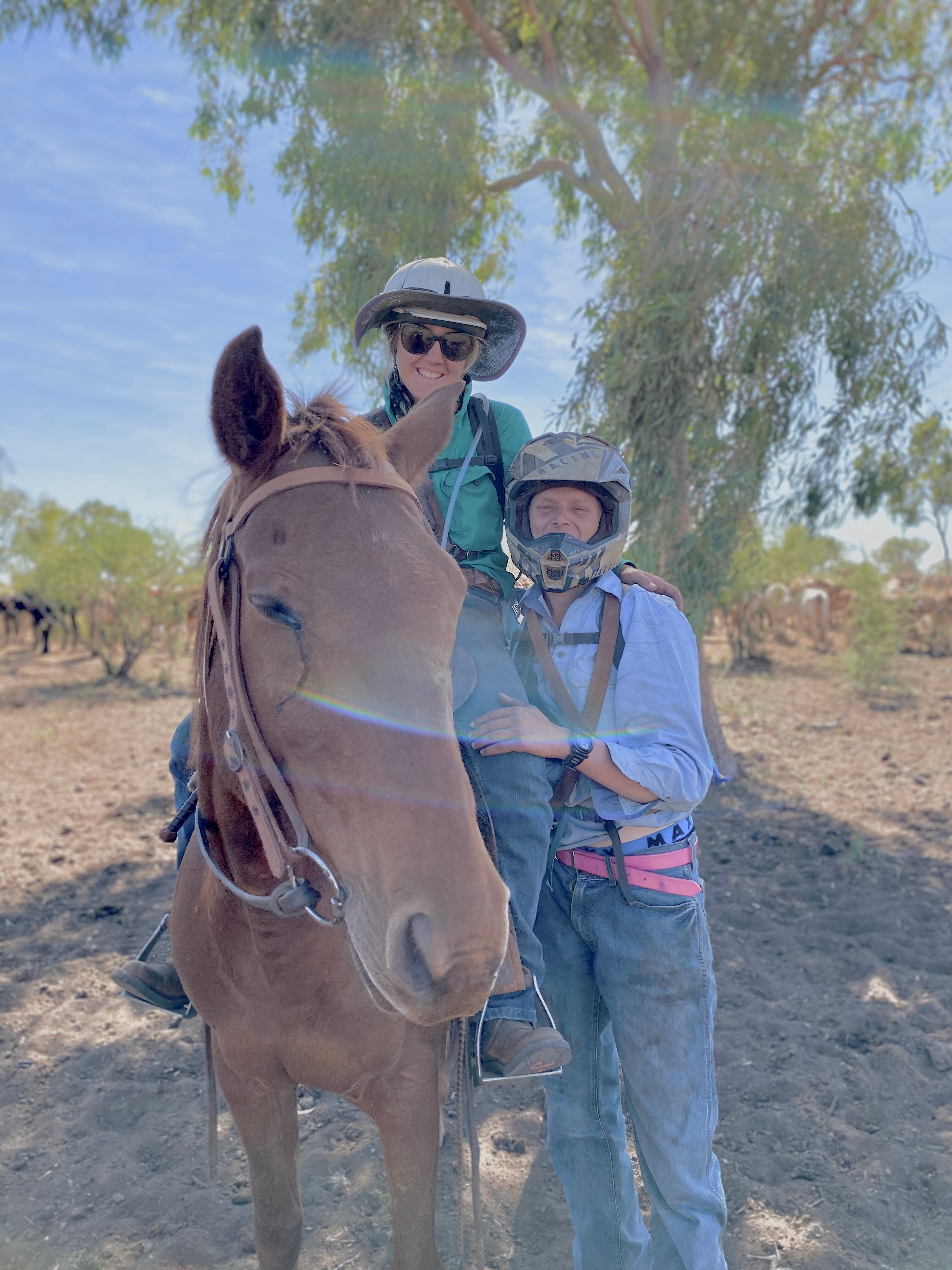 A young woman in hard hat riding a horse, a young man in a motorbike helmet stands beside them.