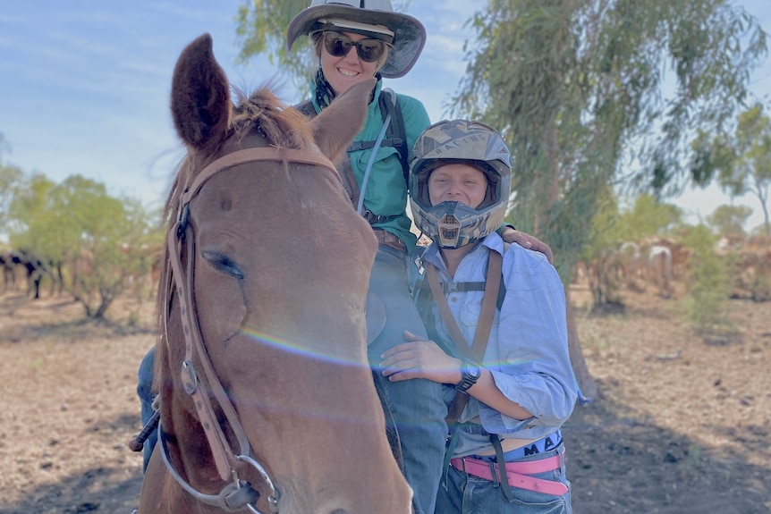 A young woman in hard hat riding a horse, a young man in motorbike helmet stands beside them.