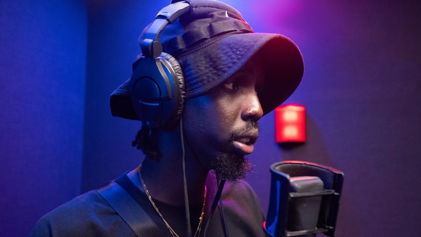 Man wearing a black hat in a studio standing in front of a microphone. A red light in the background.