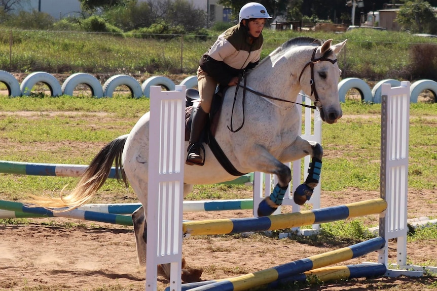 A small child rides a white horse over a small jump