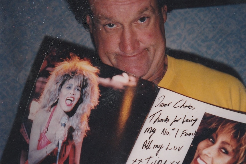 A man with curly brown hair holds a poster of singer Tina Turner which has a message written on it next to her signature.