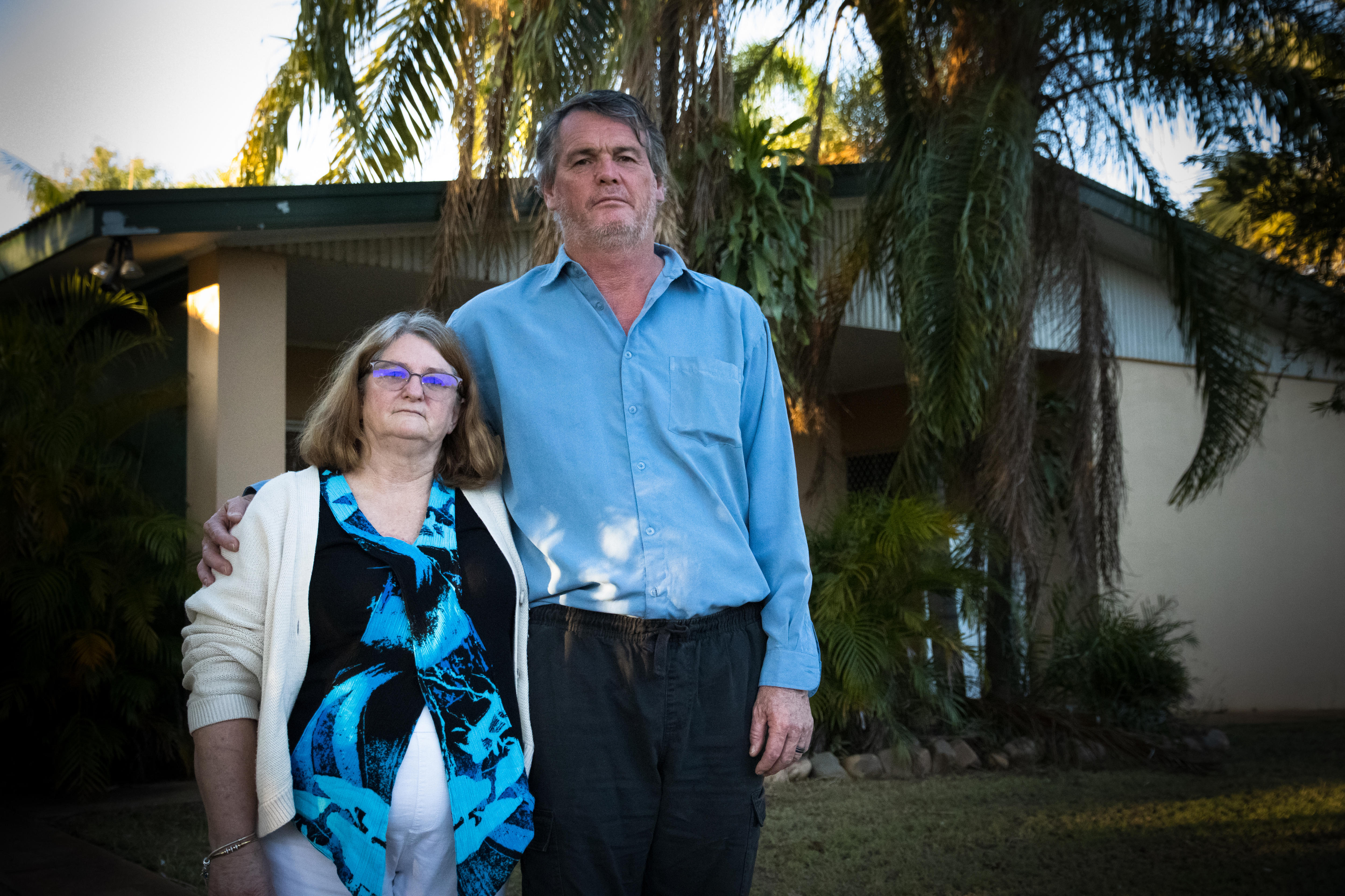 A man and woman stand side by side outside their home.  They both have a serious expression