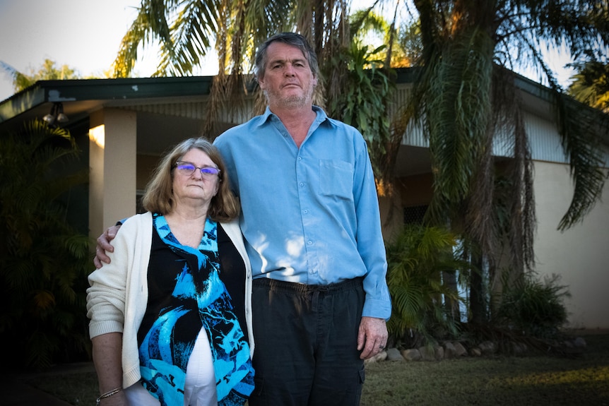 A man and woman stand side by side outside their home. They both have a serious expression