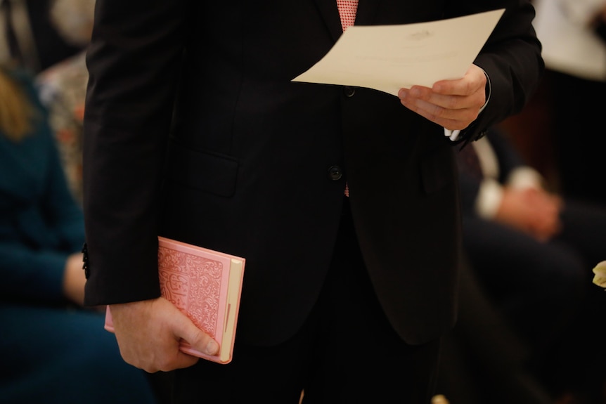 A close up shot of a man holding a Koran in one hand and a sheet of paper in the other.