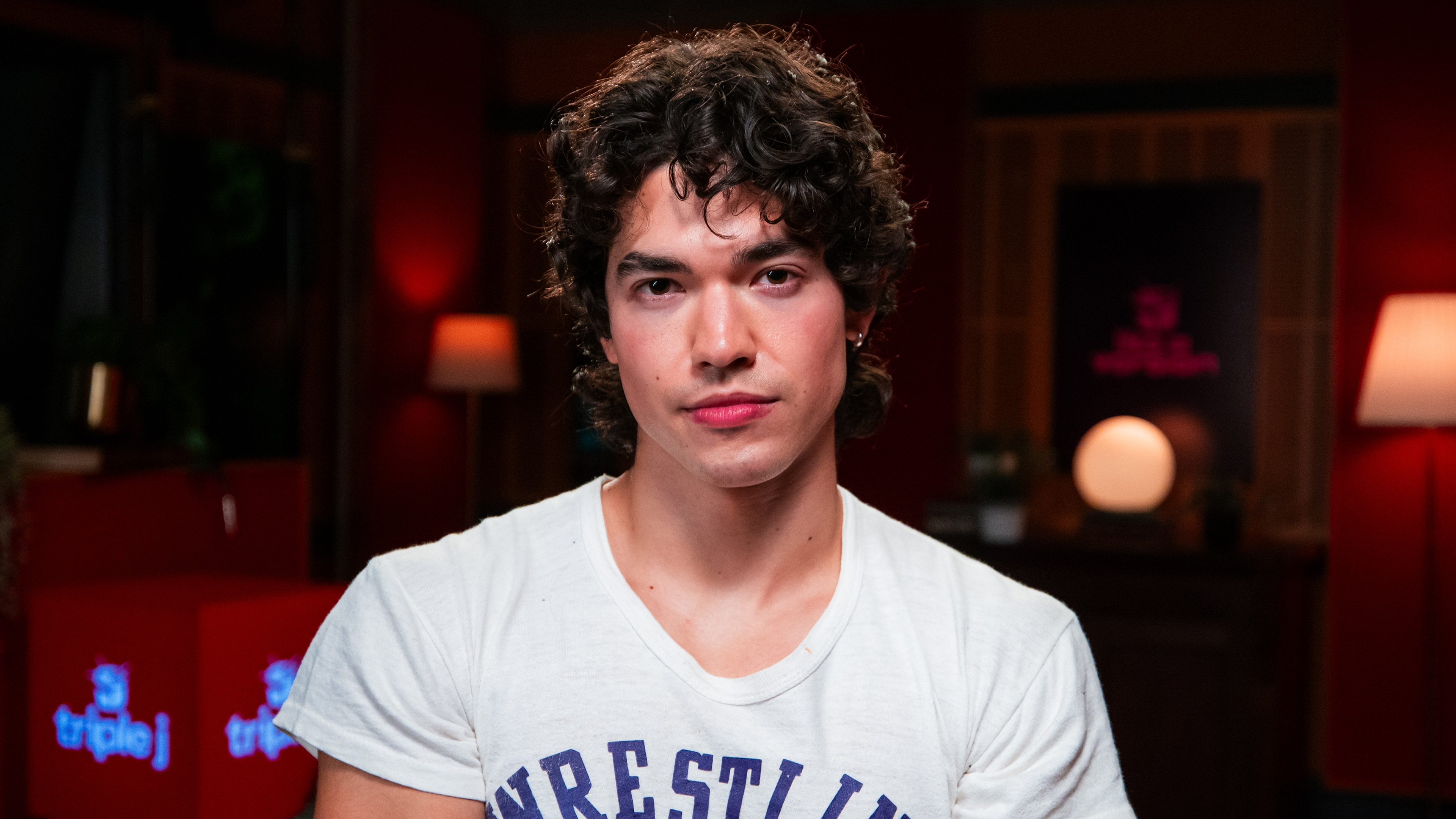 Conan Gray, wearing a white t-shirt with blue, illegible text, sits in front of a triple j lightbox.