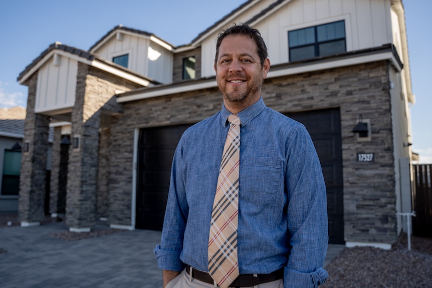 A middle-aged man wearing a blue shirt and patterned tie stands in the driveway of a grey brick home